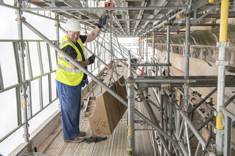 A workman standing on scaffolding hoists up stone cornicing 