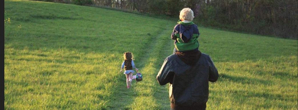 A young girl walking through a green grassy field followed by an adult carrying a toddler on their shoulders