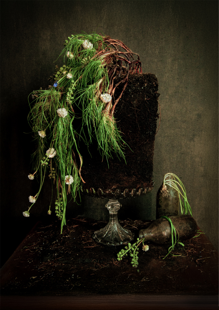 A dark still-life showing a soil cylinder draped with grasses and small white flowers