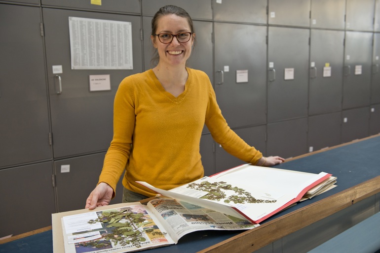 A woman smiles while holding an book open, showcasing pressed plant specimens on the pages