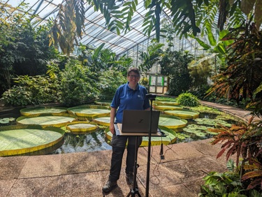 Person standing in front of a pond, surrounded by plants, inside a glasshouse.