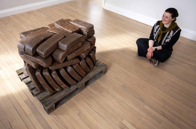 A woman sits next to a sculpture of brown ceramic blocks stacked together