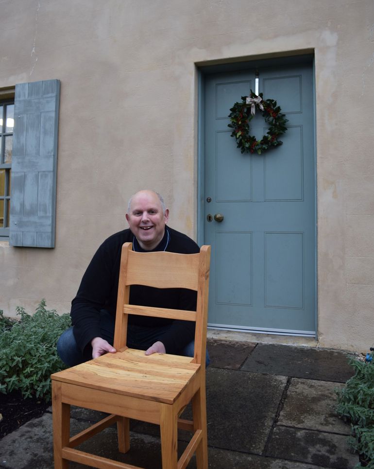 A man crouches down beside a chair in front of a door with a Christmas wreath 